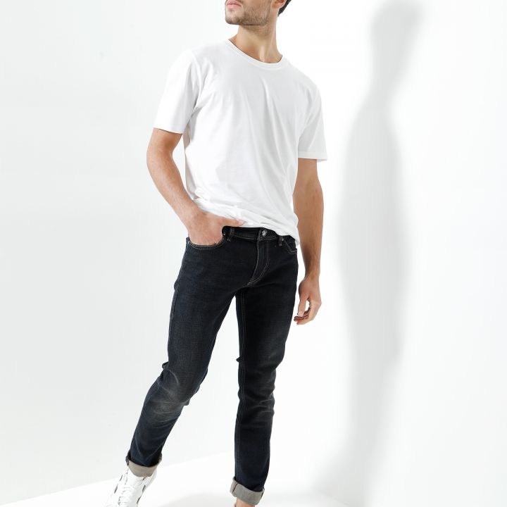 Ein Mann steht vor einem weißen Hintergrund. Er trägt ein schlichtes weißes T-Shirt, schwarze Jeans und weiße Turnschuhe. Er hat eine Hand in der Tasche und blickt mit neutralem Gesichtsausdruck zur Seite.