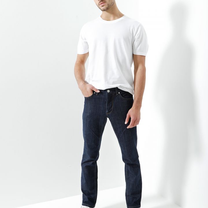 Ein Mann in weißem T-Shirt, Blue Jeans und weißen Turnschuhen steht vor einem weißen Hintergrund. Er hat eine Hand in der Tasche und nimmt eine entspannte Pose ein.