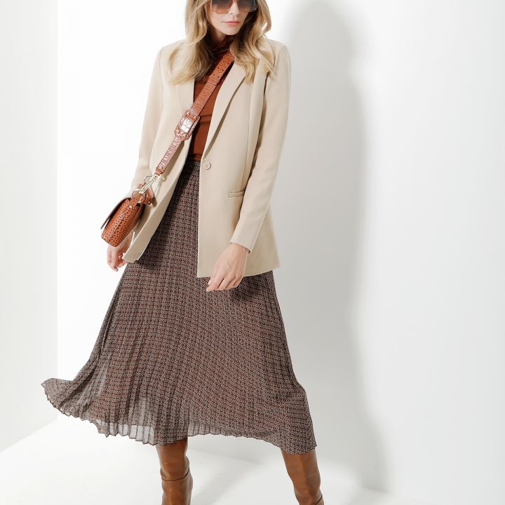 Eine Frau trägt eine Sonnenbrille und einen beigen Blazer über einem braun gemusterten Kleid. Sie trägt eine braune Umhängetasche und kniehohe braune Stiefel. Sie steht vor einem weißen Hintergrund.