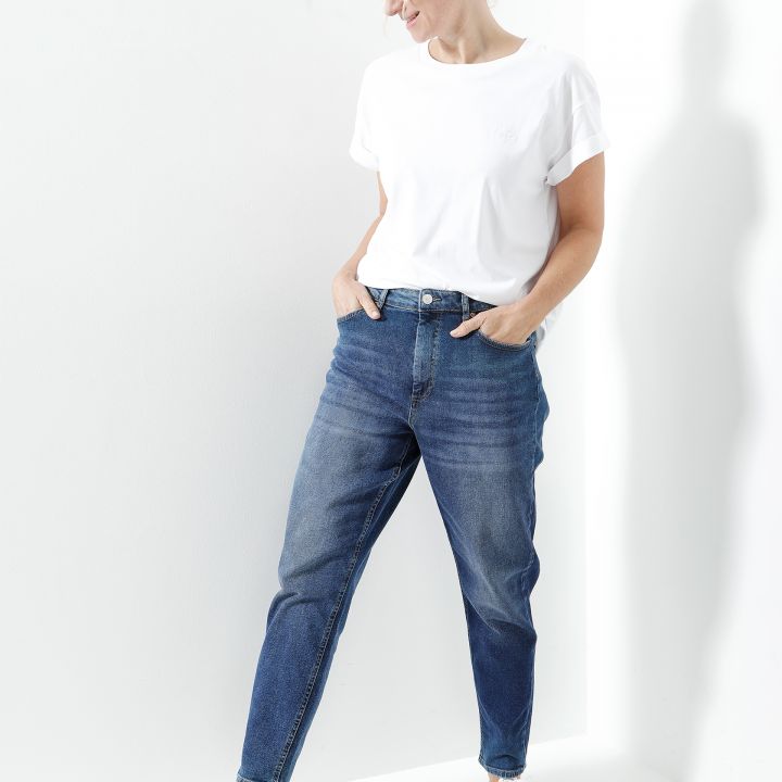Eine Person steht lässig mit den Händen in den Taschen und lehnt leicht an einer weißen Wand. Sie trägt ein weißes T-Shirt, blaue Jeans und weiße Turnschuhe. Ihr Gesichtsausdruck ist entspannt und die Umgebung ist hell mit weichen Schatten.