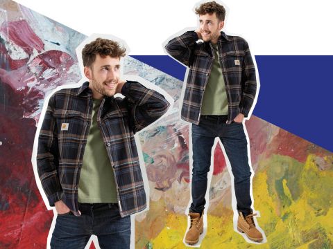 Ein Mann in kariertem Hemd und Jeans steht selbstbewusst vor einem farbenfrohen abstrakten Hintergrund. Darunter trägt er ein grünes Hemd und braune Stiefel, eine Hand hinter dem Kopf und lächelt.