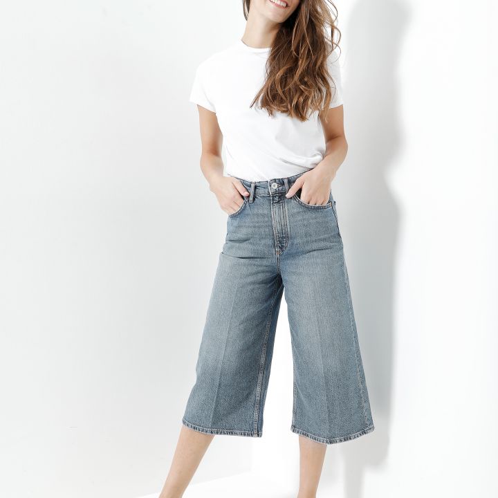 Eine Person mit langen Haaren, die ein weißes T-Shirt, weite Jeans-Culottes und weiße Turnschuhe trägt, steht vor einer weißen Wand. Sie lächelt und hat eine Hand in der Tasche.