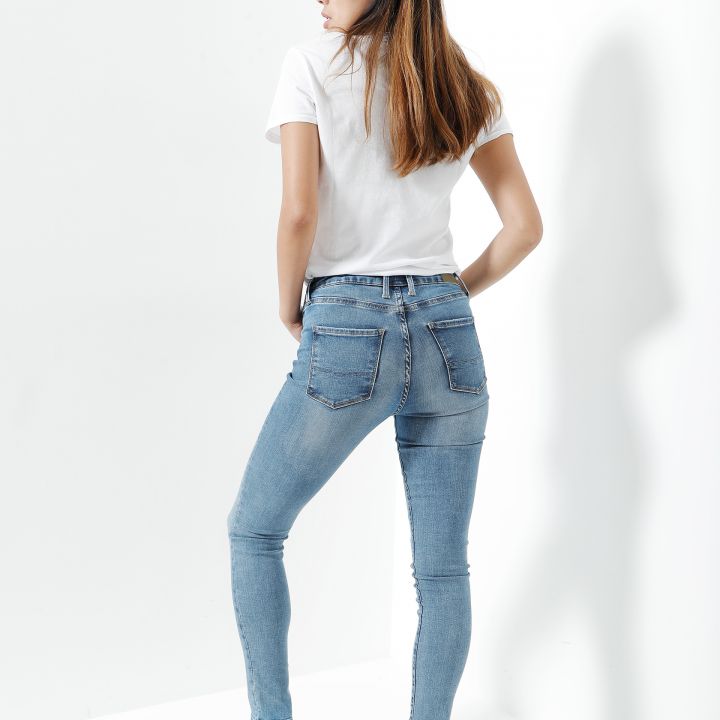 Eine Person steht mit abgewandtem Gesicht da und trägt ein weißes T-Shirt, hellblaue Jeans und weiße Turnschuhe. Der Hintergrund ist schlicht weiß und wirft einen Schatten. Die Person steht diagonal und zeigt das Outfit.