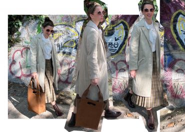 Eine Person mit Sonnenbrille und einem langen, hellen Mantel steht an einer mit Graffiti bedeckten Wand. Sie trägt ein weißes Hemd, einen karierten Rock und braune Stiefel und hält eine braune Tasche in der Hand. Das Sonnenlicht wirft weiche Schatten.