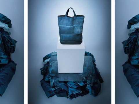 Eine blaue Einkaufstasche aus Denim steht auf einem weißen Podest, umgeben von einem Stapel verschiedener Denim-Stoffe. Das Setup steht unter blauem Licht, was eine kühle Atmosphäre schafft.