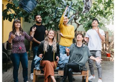 Eine Gruppe von sechs Personen steht und sitzt unter einem großen Baum. Sie lächeln und einige werfen spielerisch ihre Jacken in die Luft. Die Szene ist lebhaft und fröhlich, das Sonnenlicht fällt durch die Blätter.