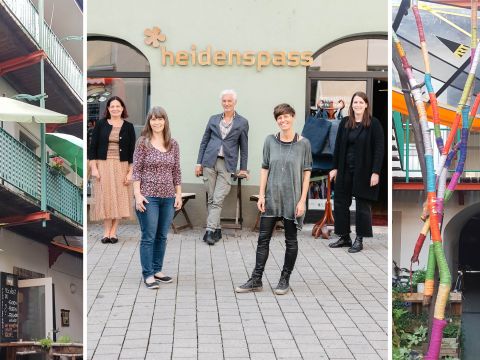 Eine Gruppe von vier Personen steht vor einem Gebäude mit einem Schild mit der Aufschrift „Heidenspass“. Sie scheinen zu lächeln und für das Foto zu posieren. Das Gebäude hat grüne Akzente und an den Seiten sind lebendige, farbenfrohe Kunstwerke zu sehen.