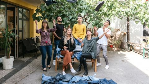 Sechs Menschen haben sich draußen versammelt und werfen fröhlich Jeans in die Luft. Sie stehen oder sitzen auf einer Bank, umgeben von Grün. Mehrere Jeans liegen ausgebreitet auf dem Boden und sorgen für eine lebendige und lockere Atmosphäre.