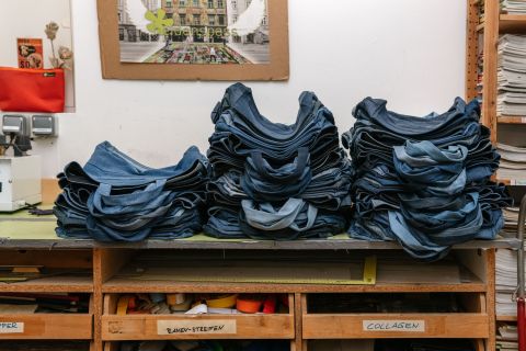 Stapel gefalteter Blue Jeans liegen auf einem Holztisch in einem Arbeitsbereich. In den Regalen darunter sind verschiedene Nähutensilien untergebracht. Im Hintergrund ist ein an der Wand befestigtes Kunstwerk zu sehen.
