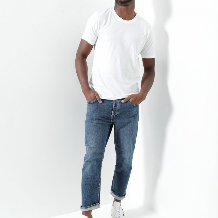 Ein Mann in weißem T-Shirt, Blue Jeans und weißen Sneakers steht vor einem weißen Hintergrund. Er hat die Hände in den Taschen und blickt mit entspannter Haltung in die Kamera.