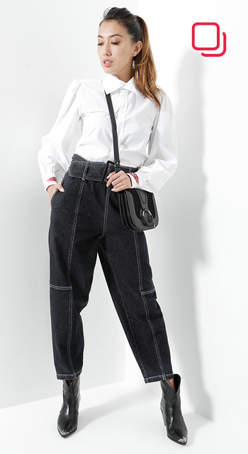 Eine Person steht vor einem weißen Hintergrund. Sie trägt eine weiße Bluse, schwarze Jeans mit hoher Taille und schwarze Stiefeletten. Sie trägt eine schwarze Umhängetasche und ihre Haare sind zurückgebunden. Das Licht wirft einen weichen Schatten.