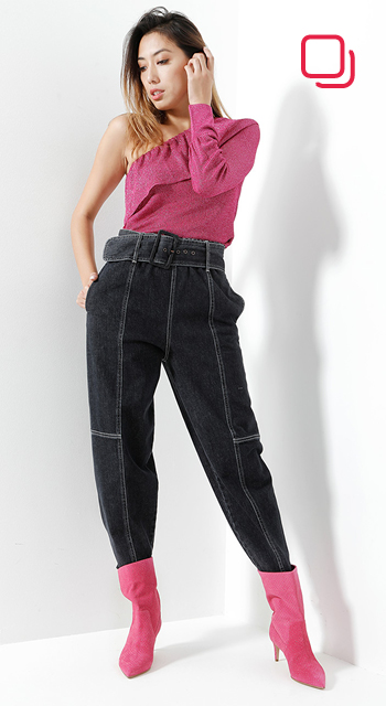 Eine Person posiert in einem stylischen Outfit: ein rosa One-Shoulder-Top, dunkle Jeans mit hoher Taille und Gürtel und rosa Stiefeletten. Sie steht vor einer weißen Wand, eine Hand steckt in der Tasche und die andere berührt ihr Haar.