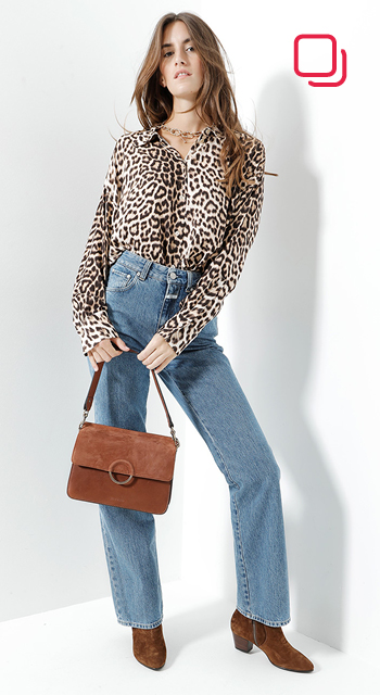 Eine Person steht selbstbewusst vor einem weißen Hintergrund und trägt eine Bluse mit Leopardenmuster, hoch taillierte Bluejeans und braune Stiefeletten. Sie hält eine braune Handtasche und hat langes, welliges Haar. In der oberen rechten Ecke ist ein rotes Logo zu sehen.