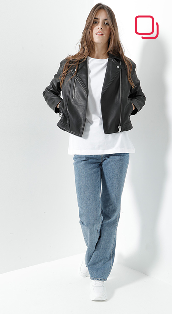 Eine Person steht vor einem weißen Hintergrund und trägt eine schwarze Lederjacke über einem weißen Hemd, blaue Jeans und weiße Turnschuhe. Sie hat lange Haare und ihre Hände stecken in den Jackentaschen. In der oberen rechten Ecke befindet sich ein rotes quadratisches Logo.