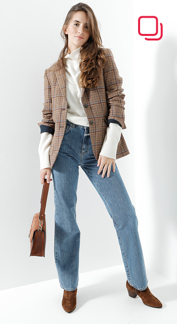 Eine Person mit langen Haaren trägt einen braun karierten Blazer über einem weißen Hemd, blaue Jeans und braune Stiefel. Sie hält eine braune Handtasche und steht vor einem weißen Hintergrund mit einem roten quadratischen Logo in der Ecke.