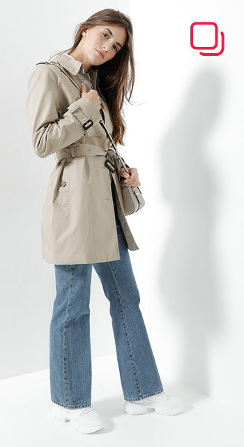 Eine Frau mit langen Haaren, einem beigen Trenchcoat und blauen Jeans, posiert mit der Hand am Kinn. Sie trägt eine kleine Umhängetasche und steht vor einem weißen Hintergrund.