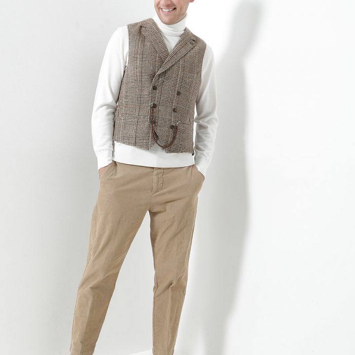 Ein lächelnder Mann steht vor einer weißen Wand. Er trägt eine graue Weste mit Hahnentrittmuster über einem weißen Rollkragenpullover, beige Hosen und braune Turnschuhe. Seine Hände stecken in den Taschen und er hat kurzes, welliges blondes Haar.