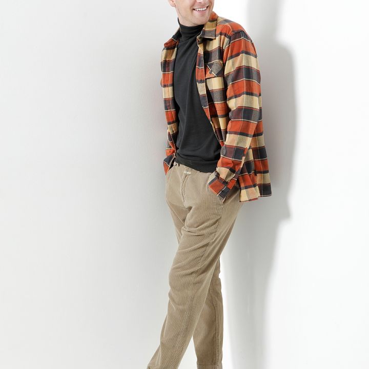 Ein Mann in schwarzem Rollkragenpullover, orange kariertem Hemd und beiger Hose lehnt lächelnd an einer weißen Wand. Er trägt braune Schuhe und hat die Hände in den Taschen.