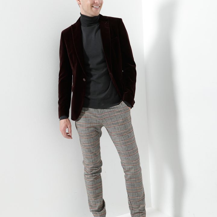 Ein Mann steht lächelnd in einem gut beleuchteten Raum. Er trägt eine dunkle Samtjacke, einen schwarzen Rollkragenpullover, karierte Hosen und schwarze Schuhe. Er lehnt leicht an einer weißen Wand und strahlt ein selbstbewusstes und stilvolles Auftreten aus.