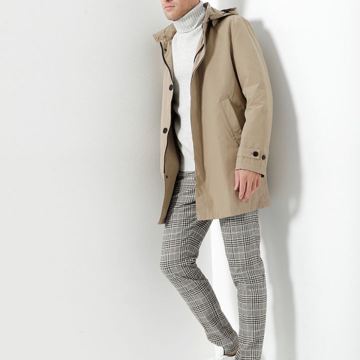 Eine Person, die einen beigen Mantel über einem weißen Rollkragenpullover, karierte Hosen und weiße Turnschuhe trägt, lehnt an einer weißen Wand. Die Person blickt in einer hellen, minimalistischen Umgebung zur Seite.