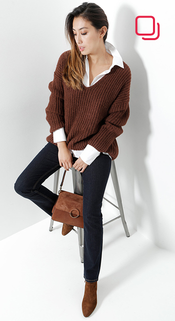 Eine Person in einem braunen Pullover mit weißem Hemd darunter und dunkler Jeans sitzt auf einem Hocker. Sie hält eine braune Handtasche und trägt braune Stiefeletten. Der Hintergrund ist weiß.