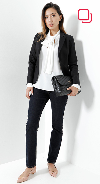 Eine Frau steht vor einem weißen Hintergrund und trägt einen schwarzen Blazer, eine weiße Bluse mit Schleife und dunkle Jeans. Sie hält eine schwarze gesteppte Clutch unter dem Arm und trägt Schuhe in neutralen Farben. In der oberen rechten Ecke befindet sich ein rotes Logo.
