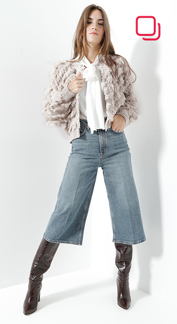 Eine Person posiert selbstbewusst in einer strukturierten beigefarbenen Strickjacke, einer weißen Bluse mit Schleife, Jeans mit weitem Bein und hohen braunen Stiefeln. Vor einem weißen Hintergrund wirken sie modisch.