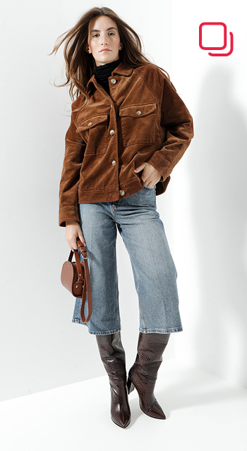 Eine Person posiert in einer braunen Wildlederjacke und blauen Jeans und hält eine braune Handtasche. Sie trägt hohe braune Stiefel und steht vor einem weißen Hintergrund. In der oberen rechten Ecke befindet sich ein rotes Logo mit einem Quadrat und einem Kreis.