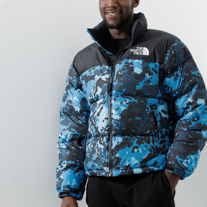 Eine Person, die eine Pufferjacke mit blauem und schwarzem Camouflage-Muster trägt, steht lächelnd vor einem einfarbigen Hintergrund. Auf der Brust der Jacke ist das Logo von The North Face zu sehen. Die Person trägt eine schwarze Hose.