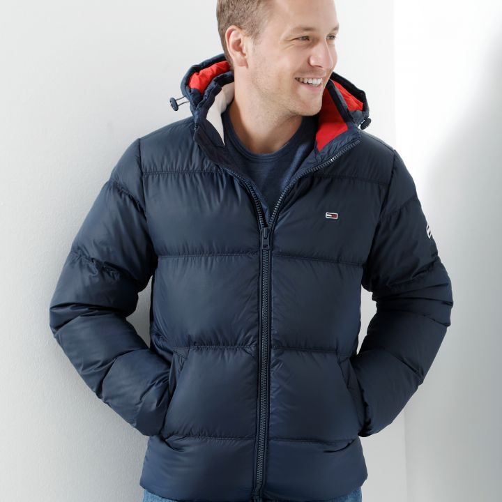 Eine Person in einer marineblauen Pufferjacke mit rotem Futter und blauen Jeans steht lächelnd vor einer weißen Wand und blickt zur Seite. Die Hände stecken in den Jackentaschen.