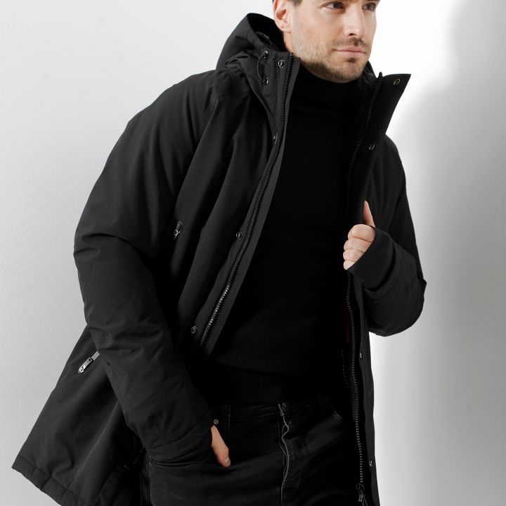 Ein Mann in schwarzer Jacke und schwarzem Rollkragen steht vor einem neutralen Hintergrund. Er hat kurzes dunkles Haar und einen Bart, seine linke Hand steckt in der Tasche und mit der rechten richtet er seine Jacke.
