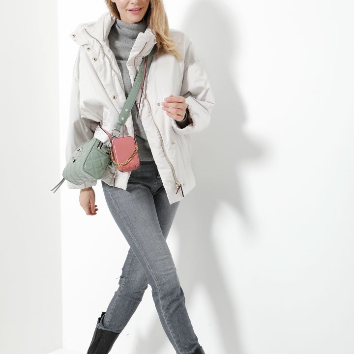 Eine Frau in weißer Jacke und grauen Jeans geht selbstbewusst. Sie trägt eine grüne Umhängetasche und schwarze Stiefel. Der Hintergrund ist schlicht und gut beleuchtet und wirft einen Schatten neben sie.