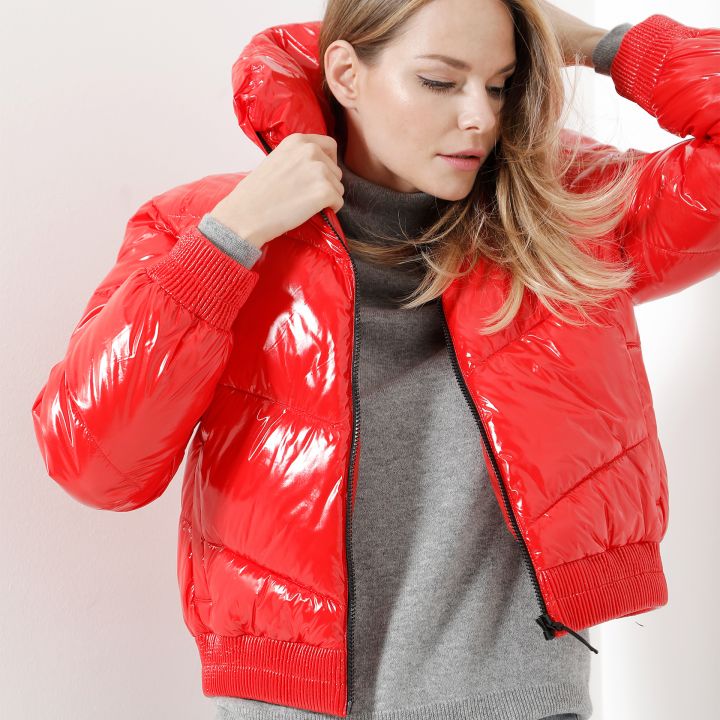 Eine Person, die eine glänzende rote Pufferjacke über einem grauen Pullover trägt, rückt die Kapuze zurecht. Sie hat lange Haare und steht vor einem weißen Hintergrund.
