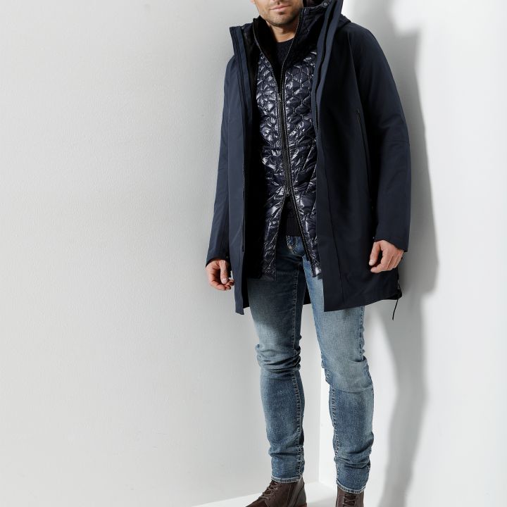Ein Mann steht vor einer weißen Wand. Er trägt eine marineblaue Jacke über einer glänzenden Steppweste, blaue Jeans und braune Lederstiefel. Er blickt mit neutralem Gesichtsausdruck in die Kamera.