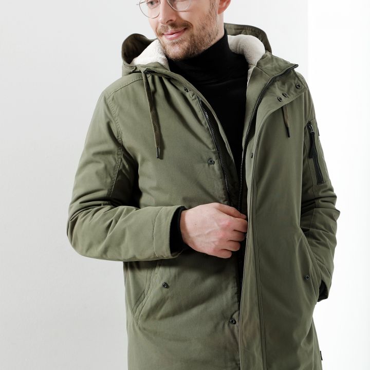 Ein Mann trägt eine Brille und eine grüne Parka mit Kapuze und Fleecefutter. Er hat einen leichten Bart und blickt zur Seite, während er mit einer Hand die Vorderseite seiner Jacke zurechtrückt. Der Hintergrund ist schlicht und weiß.