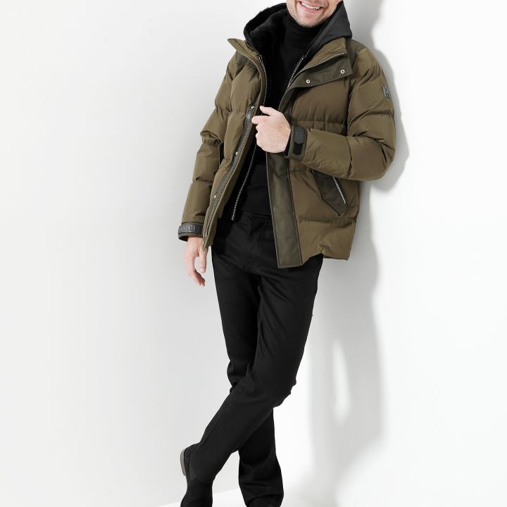 Eine Person lehnt lächelnd an einer weißen Wand. Sie trägt eine olivgrüne Pufferjacke, schwarze Hosen und schwarze Schuhe. Das Outfit wird durch einen schwarzen Rollkragenpullover ergänzt und eine Hand ruht in einer Tasche.