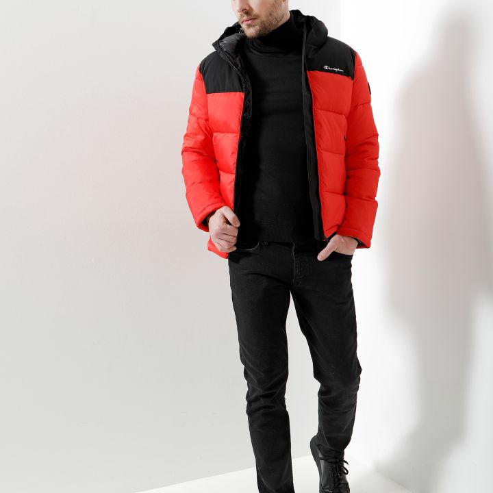 Ein Mann in einer rot-schwarzen Pufferjacke und schwarzen Hosen läuft vor einem weißen Hintergrund. Sein Kopf ist leicht geneigt, während er nach unten blickt. Seine braunen Haare sind lässig gestylt und er trägt schwarze Turnschuhe.