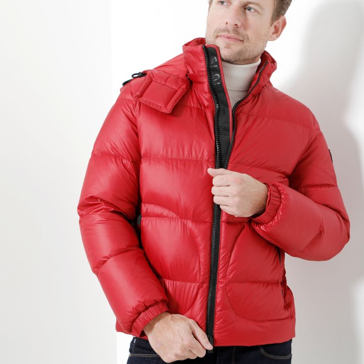 Ein Mann, der eine leuchtend rote Pufferjacke über einem weißen Rollkragenpullover trägt, steht vor einem weißen Hintergrund. Er hält mit einer Hand den Reißverschluss der Jacke und blickt zur Seite. Außerdem trägt er dunkle Jeans.