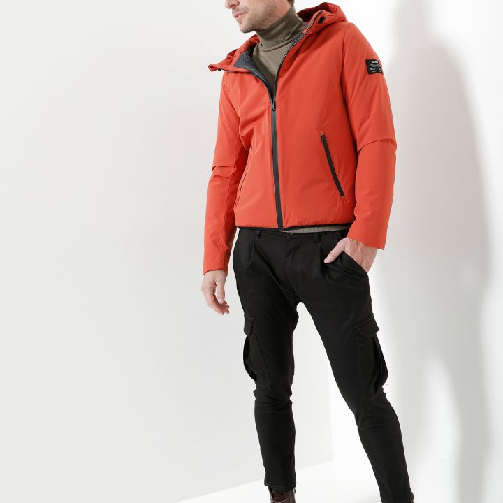 Ein Mann in einer roten Kapuzenjacke und einem olivgrünen Rollkragenpullover steht vor einem weißen Hintergrund. Er trägt schwarze Hosen und braune Stiefel, blickt nach unten und zur Seite und hat die Hände in den Taschen.