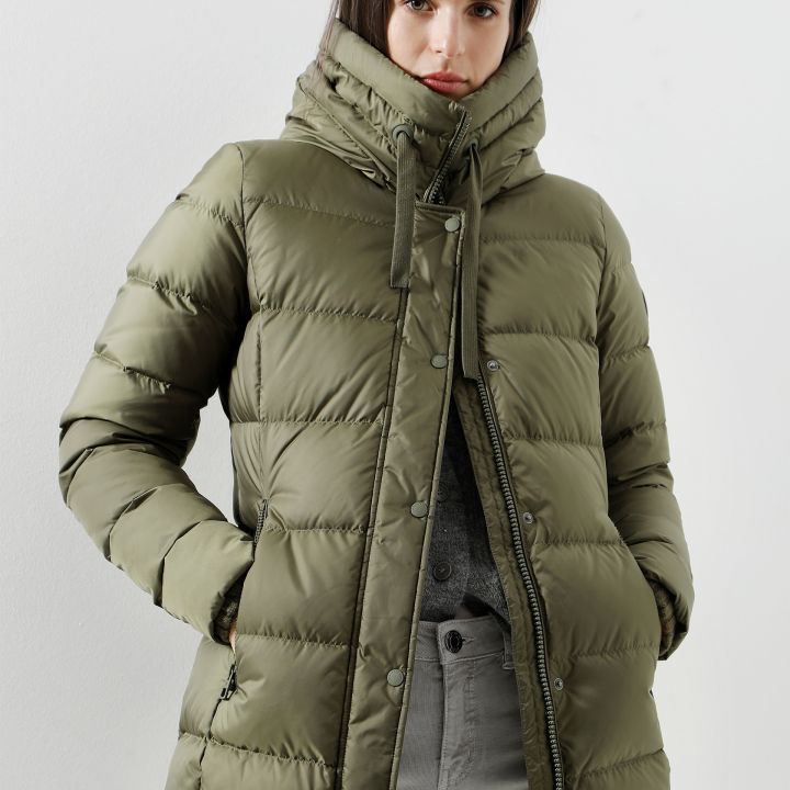 Eine Person in einer olivgrünen Pufferjacke steht vor einem schlichten weißen Hintergrund. Die Jacke hat einen hohen Kragen und Reißverschlüsse. Die Person hat die Hände in den Taschen und trägt hellgraue Hosen.