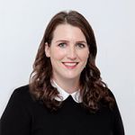 Eine Frau mit langen braunen Haaren und einer weißen Bluse unter einem schwarzen Pullover lächelt vor einem schlichten weißen Hintergrund in die Kamera.