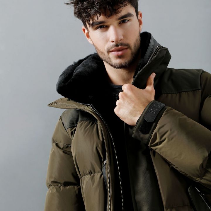 Mann mit lockigem Haar, trägt eine grüne Pufferjacke mit Kapuze und blickt vor neutralem Hintergrund in die Kamera.