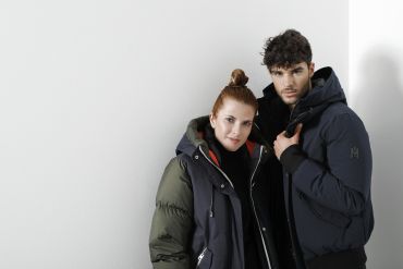 Eine Person mit hochgesteckten langen Haaren trägt eine olivgrüne und schwarze Jacke. Daneben trägt eine Person mit lockigem Haar eine dunkelblaue Jacke. Beide stehen vor einem weißen Hintergrund und blicken in die Kamera.