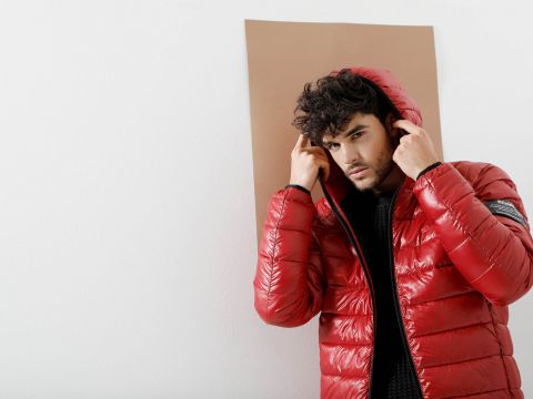 Eine Person in einer glänzenden roten Pufferjacke mit hochgezogener Kapuze steht vor einem schlichten Hintergrund. Sie scheint die Kapuze mit den Händen zurechtzurücken und blickt dabei aufmerksam in die Kamera.