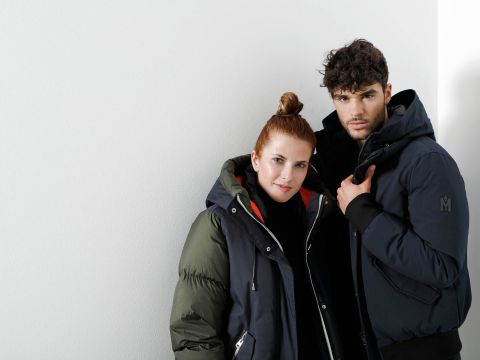 Eine Frau und ein Mann stehen vor einer weißen Wand. Beide tragen dunkle Winterjacken. Die Frau hat einen Dutt und der Mann lockiges Haar. Sie blicken mit ruhigem Gesichtsausdruck direkt in die Kamera.