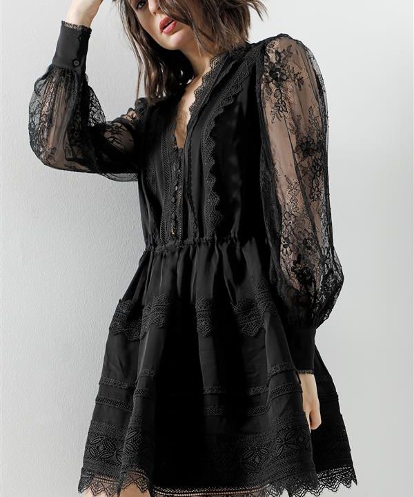 Eine Frau in einem eleganten schwarzen Kleid mit Spitzendetails an Ärmeln und Mieder steht vor einem hellen Hintergrund. Das Kleid hat lange, transparente Ärmel und einen kurzen Rock mit aufwendigen Spitzenmustern.