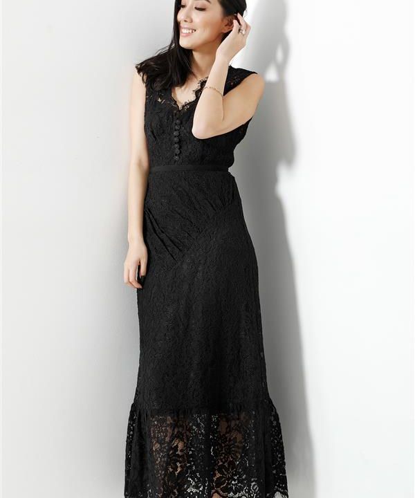 Eine Frau in einem schwarzen Spitzenkleid steht vor einer weißen Wand, lächelt und berührt ihr Haar. Das Kleid ist ärmellos, hat eine taillierte Taille und einen durchsichtigen Spitzensaum. Die Beleuchtung erzeugt weiche Schatten hinter ihr.