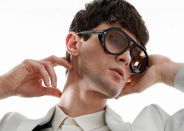 Eine Person mit kurzen dunklen Haaren trägt eine übergroße Sonnenbrille und einen hellen Anzug. Sie hat ihre Hände nahe am Hals positioniert und ihren Kopf leicht nach hinten geneigt. Der Hintergrund ist schlicht weiß.