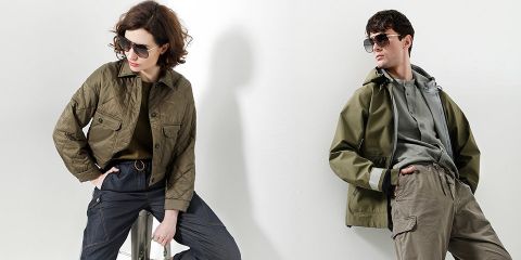 Zwei Personen in stylischer Oberbekleidung und Sonnenbrillen posieren in einem gut beleuchteten Studio. Die Person links trägt eine olivfarbene Steppjacke und dunkle Hosen, während die Person rechts eine olivfarbene Kapuzenjacke und Cargohosen trägt. Beide strahlen eine selbstbewusste, urbane Ausstrahlung aus.