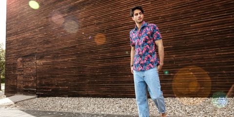 Eine Person steht im Freien und trägt ein buntes Hemd mit Blumenmuster und hellblaue Jeans. Sie steht vor einer Holzwand, unter der sich ein Kiesweg befindet. Sonnenlicht erzeugt im Bild Linsenreflexionen.
